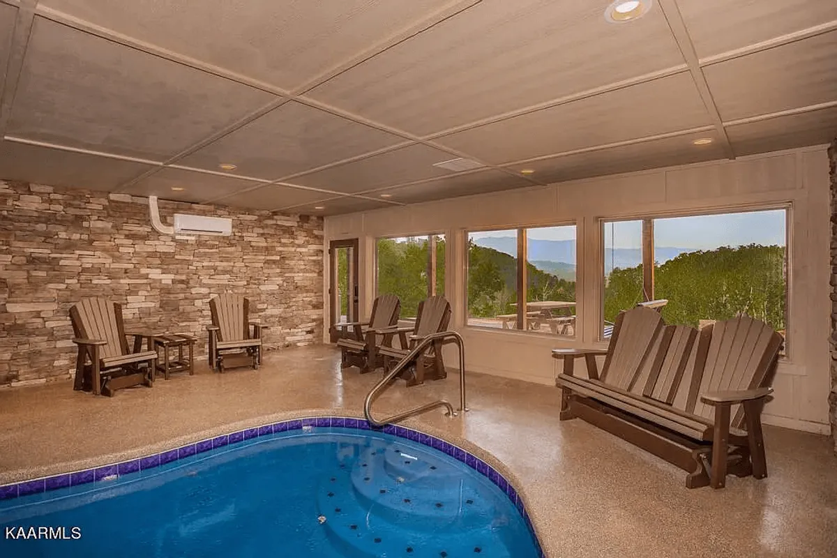 A luxurious indoor pool area features a serene swimming pool, flanked by large windows that flood the space with natural light. Stone walls and wooden chairs create a calming, resort-like atmosphere where guests can relax, socialize, or enjoy a drink poolside while taking in scenic views.