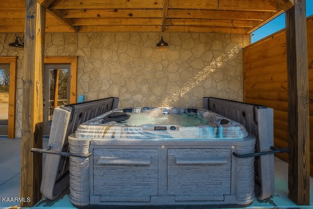 A private outdoor hot tub set beneath wooden beams and beside a rustic stone wall, with the surrounding mountain landscape visible. The scene invites relaxation and rejuvenation after an adventurous day outdoors.