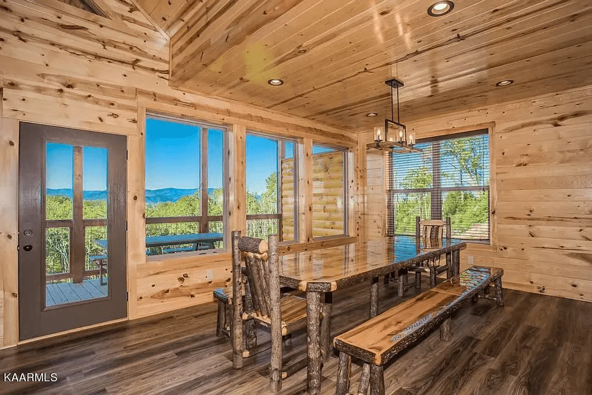 A generous wooden dining table surrounded by chairs sits beside floor-to-ceiling windows that frame stunning mountain vistas. The space is set for communal meals, games, and shared moments, with natural light pouring in to create a bright, inviting ambiance.