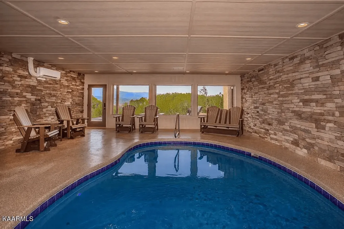 An interior scene featuring a sparkling indoor pool surrounded by large windows, adirondack chairs, and a rustic stone wall. The tranquil setting hints at both energetic play and restful relaxation, perfect for families looking to continue the fun indoors.