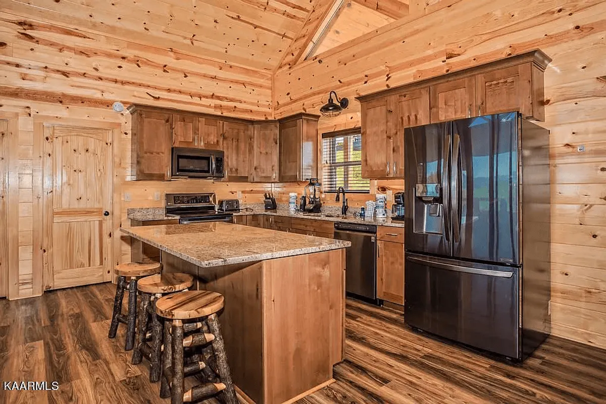 This image features a modern kitchen with a granite countertop island, wooden cabinetry, stools, and up-to-date appliances. The space is brightly lit and arranged to encourage gathering, cooking, and casual dining, epitomizing a chef’s kitchen and a social hub.