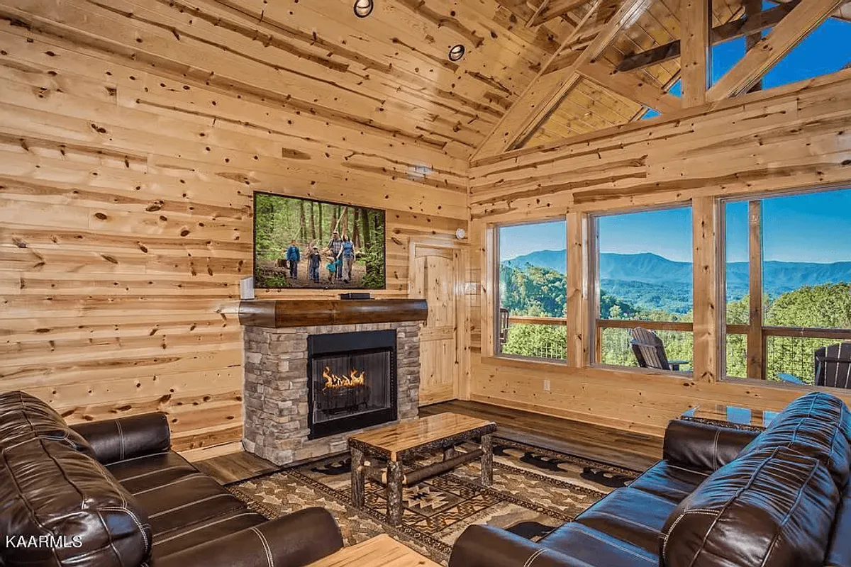 A welcoming living room with a cozy fireplace as its centerpiece, large windows letting in natural light, a comfortable sofa, and a television. Outside the windows, beautiful mountain views can be glimpsed, connecting the interior with the surrounding landscape.