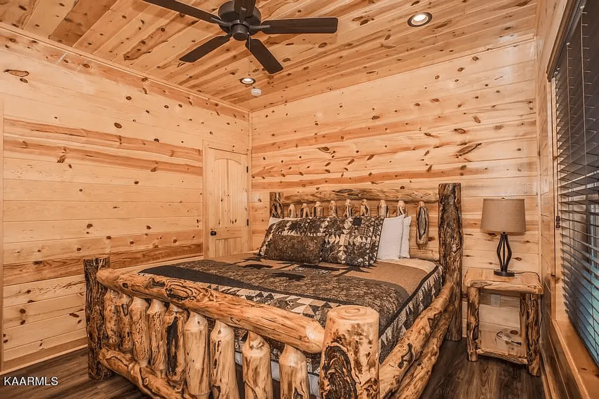 A warmly lit bedroom featuring a rustic log bed with decorative pillows, wooden walls, and a cozy nightstand. The inviting atmosphere is enhanced by natural light streaming in through nearby windows.